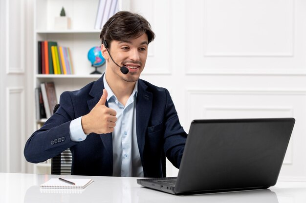 Chico guapo de servicio al cliente con auriculares y computadora portátil en traje que muestra un buen gesto