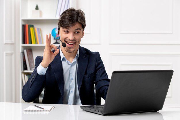 Chico guapo de servicio al cliente con auriculares y computadora portátil en traje que muestra un gesto correcto