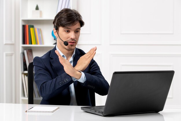 Chico guapo de servicio al cliente con auriculares y computadora portátil en traje que muestra gesto de parada