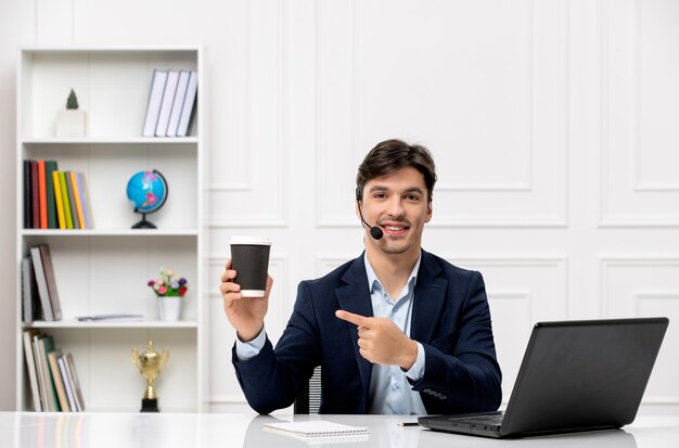 Chico guapo de servicio al cliente con auriculares y computadora portátil en traje sonriendo y sosteniendo una taza de papel