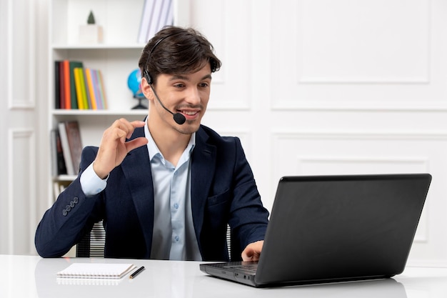 Chico guapo de servicio al cliente con auriculares y computadora portátil en traje sonriendo en videollamada