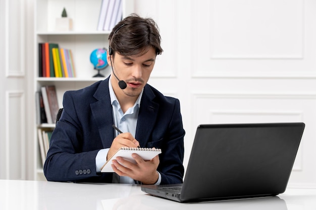 Chico guapo de servicio al cliente con auriculares y computadora portátil en traje tomando notas