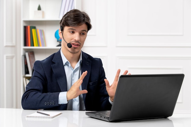 Chico guapo de servicio al cliente con auriculares y computadora portátil en traje con videollamada seria