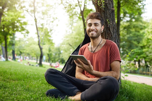 Chico guapo se sienta en la hierba del parque, lee un libro de tableta digital, conecta wifi y mira las redes sociales