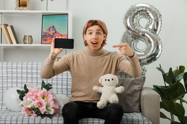 Chico guapo sonriente en el día de la mujer feliz sosteniendo y apunta al oso de peluche con el teléfono sentado en el sofá en la sala de estar