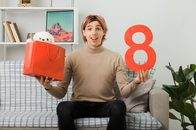 Chico guapo sonriente en el día de la mujer feliz sosteniendo el número ocho con bolsa de regalo sentado en el sofá en la sala de estar