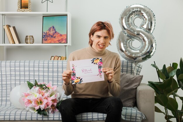 Chico guapo sonriente en el día de la mujer feliz con tarjeta de felicitación sentado en el sofá en la sala de estar