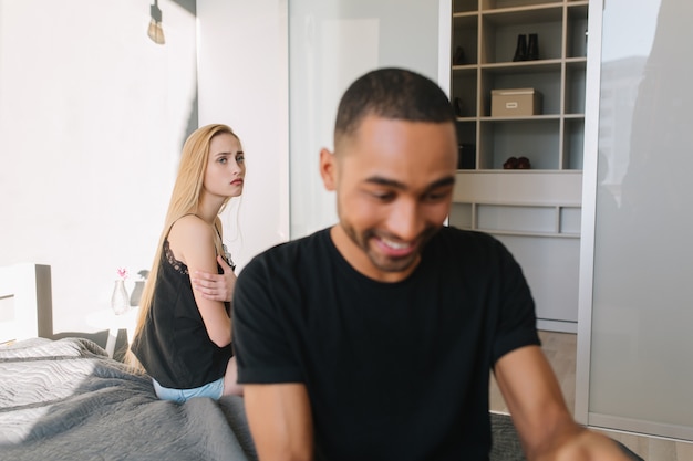 Chico guapo sonriente emocionado está ocupado con la tableta en la cama. Mujer bonita joven solitaria trastornada con el pelo rubio largo que lo mira. Divirtiéndose, relación, en casa, soledad