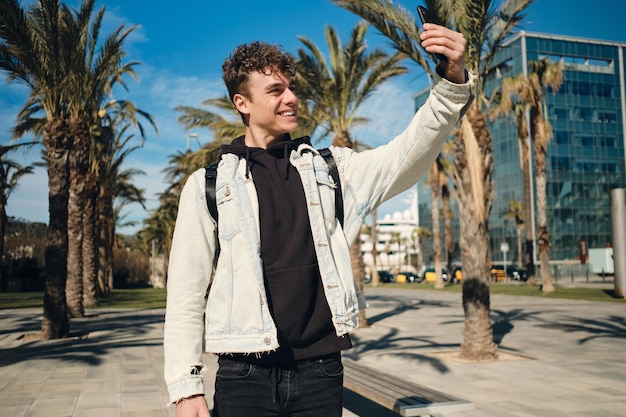 Chico guapo sonriente felizmente tomando selfie en el teléfono inteligente en el parque de la ciudad. Joven turista con estilo caminando al aire libre
