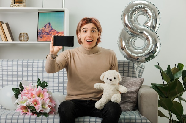 Chico guapo sorprendido en el día de la mujer feliz con oso de peluche con teléfono sentado en el sofá en la sala de estar