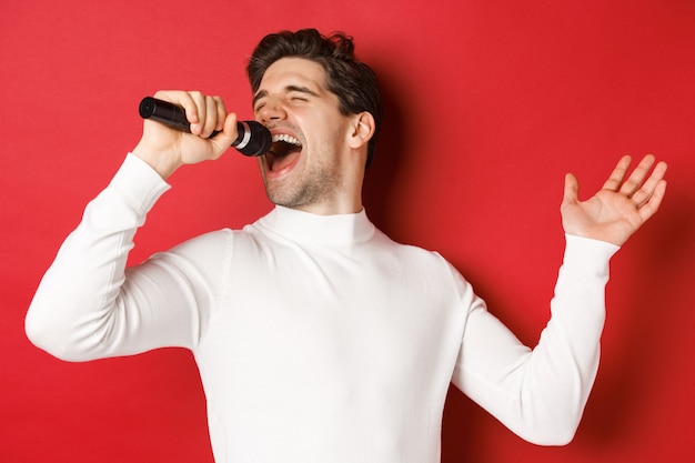 Foto gratuita chico guapo en suéter blanco, cantando una canción, sosteniendo el micrófono y actuando en el bar de karaoke, de pie sobre un fondo rojo.