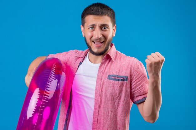 Chico guapo viajero joven con anillo inflable levantando el puño salido y feliz ganador concepto de pie sobre fondo azul.