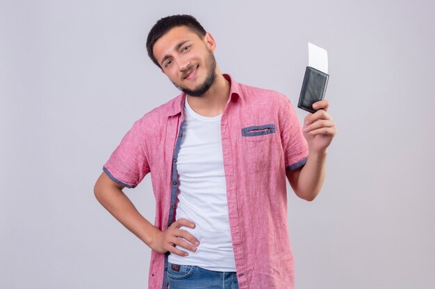 Chico guapo viajero joven con billetes de avión mirando a la cámara con una sonrisa segura de pie positivo y feliz sobre fondo blanco