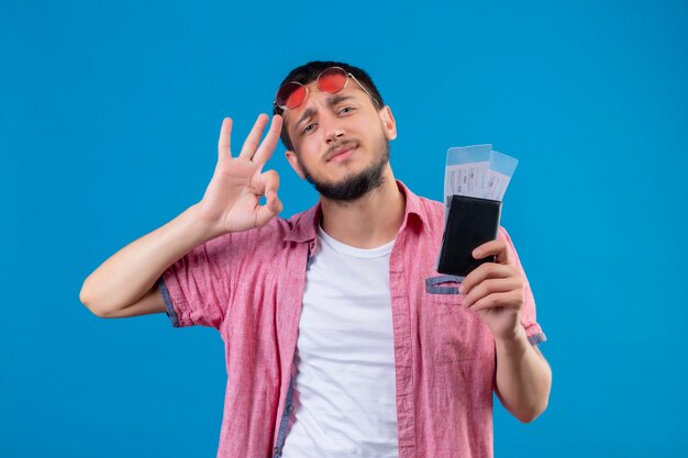 Chico guapo viajero joven con billetes de avión mirando seguro haciendo bien firmar de pie sobre fondo azul.