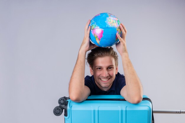 Chico guapo viajero joven de pie con maleta sosteniendo globo mirando a la cámara sonriendo alegremente con cara feliz sobre fondo blanco