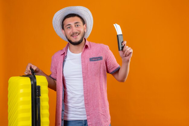 Chico guapo viajero joven con sombrero de verano con maleta y boletos aéreos mirando seguro de sí mismo sonriendo alegremente listo para viajar de pie sobre fondo naranja