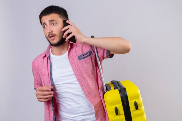 Chico guapo viajero joven vistiendo sosteniendo la maleta hablando por teléfono móvil mirando confundido de pie sobre fondo blanco.