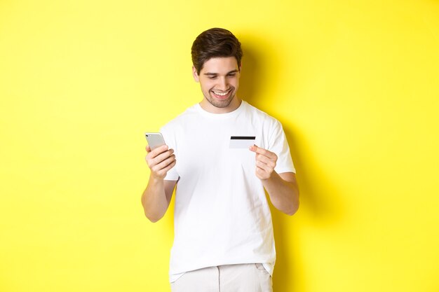 Chico haciendo un pedido en línea, registra la tarjeta de crédito en la aplicación móvil, sosteniendo el teléfono inteligente y sonriendo, de pie sobre fondo amarillo