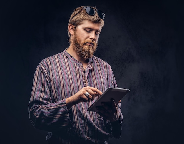 Chico hipster barbudo pelirrojo vestido con una camisa pasada de moda usando una tableta. Aislado en un fondo oscuro.