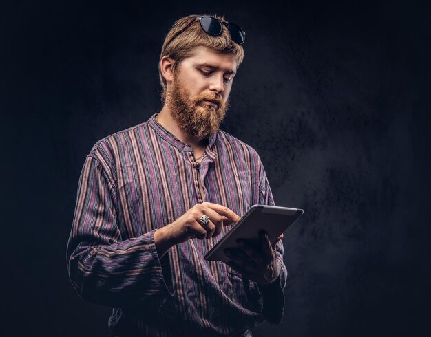 Chico hipster barbudo pelirrojo vestido con una camisa pasada de moda usando una tableta. Aislado en un fondo oscuro.