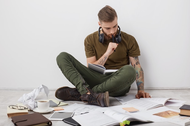 Chico hipster concentrado con tatuajes, se sienta con las piernas cruzadas en el suelo, lee libros y escribe notas