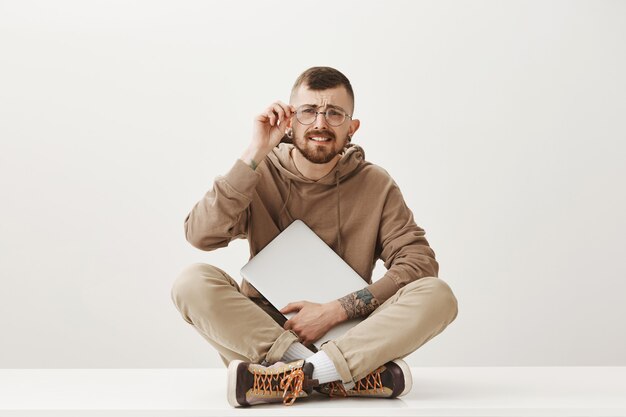 Chico hipster confundido sentado con las piernas cruzadas con la computadora portátil, ponerse gafas para ver