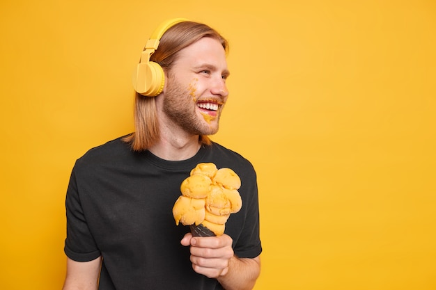 Chico hipster despreocupado con cabello pelirrojo sonríe enfocado positivamente se divierte come helado sabroso tiene cara sucia escucha música a través de auriculares posa contra el espacio de copia de la pared amarilla a la derecha