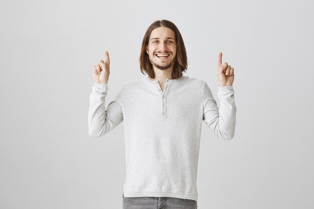 Chico hipster emocionado apuntando con el dedo hacia arriba y sonriendo