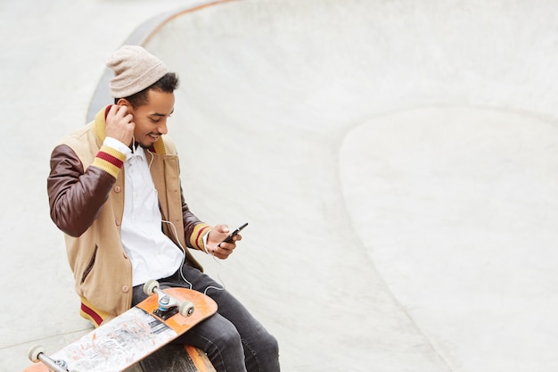 Foto gratuita chico hipster con estilo urbano se sienta cerca de su patineta, viste ropa de moda,