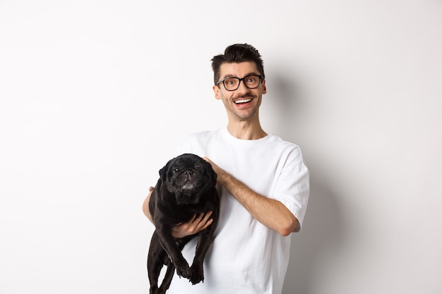 Chico hipster feliz en gafas perro mascota y sonriendo. Lindo pug negro disfruta pasar tiempo con el propietario, mirando satisfecho, de pie sobre fondo blanco.