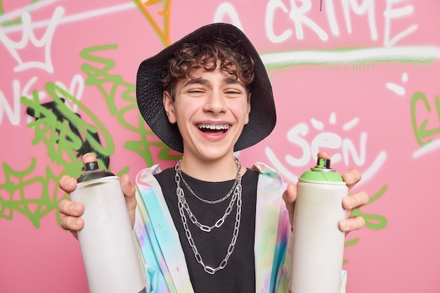 Foto gratuita el chico hipster rizado positivo se ríe y se divierte con un amigo mientras dibuja grafitis sonríe ampliamente tiene frenillos en los dientes sostiene botellas de aerosol usa ropa elegante se siente frío