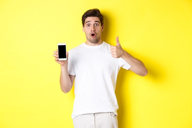 Chico impresionado que muestra una aplicación de teléfono inteligente impresionante en la pantalla y el pulgar hacia arriba, jadeando asombrado, de pie