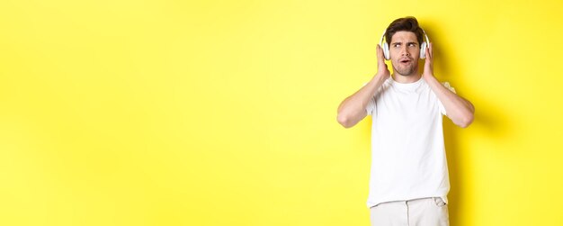 Foto gratuita chico intrigado disfrutando de melodías en auriculares escuchando atentamente música en auriculares de pie sobre amarillo