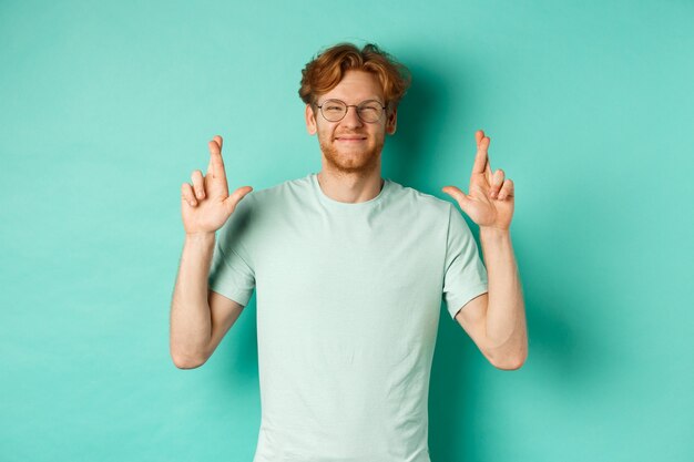 Chico joven alegre con cabello rojo y barba, con gafas, sonriendo y cruzando los dedos para la buena suerte, pidiendo deseos y luciendo optimista, de pie sobre fondo de menta