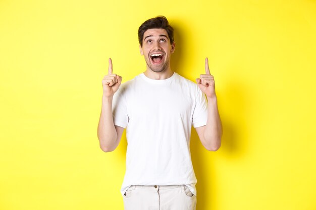 Chico joven alegre en camiseta blanca reaccionando a la oferta promocional, apuntando y mirando hacia arriba con asombro, de pie sobre fondo amarillo.