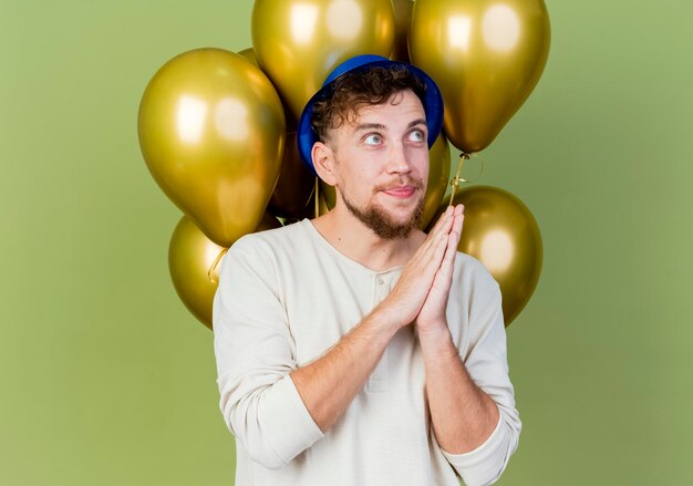 Chico joven apuesto partido eslavo con sombrero de fiesta de pie delante de globos manteniendo las manos juntas mirando al lado pidiendo algo aislado en la pared verde oliva