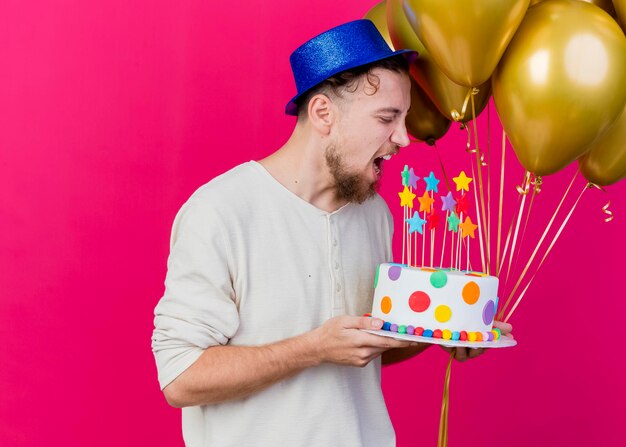 Chico joven apuesto partido eslavo con sombrero de fiesta sosteniendo globos y pastel de cumpleaños con estrellas preparándose para morder el pastel aislado en la pared rosa con espacio de copia