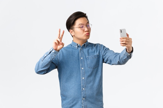 Chico joven asiático lindo y divertido haciendo pucheros tontamente, tomando selfie en teléfono inteligente, usando la aplicación de filtro de fotos para cambiar de apariencia, disparándose a sí mismo con el signo de la paz y un beso, fondo blanco.