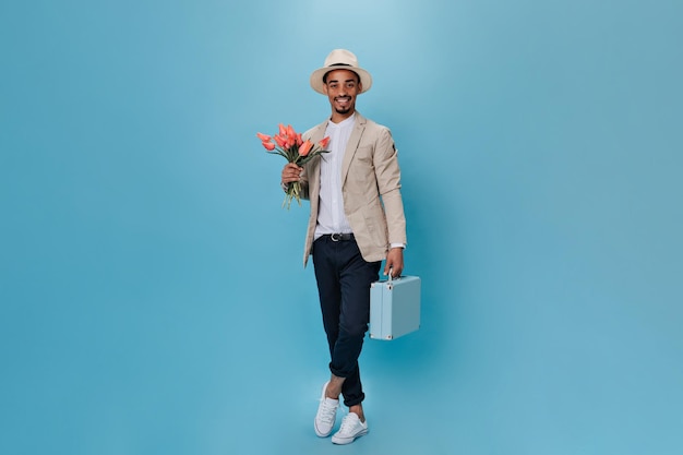 Chico joven atractivo con sombrero posando con flores rosas y maleta Instantánea completa del hombre con chaqueta y pantalón negro sosteniendo tulipanes sobre fondo aislado