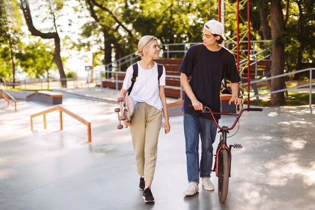 Chico joven con bicicleta y chica guapa con monopatín felizmente pasando tiempo juntos en el skatepark moderno