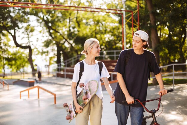 Chico joven con bicicleta y una chica muy sonriente con patineta pasando tiempo juntos en el skatepark moderno