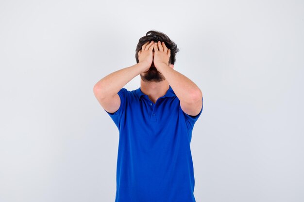 Chico joven en camiseta azul que cubre parte de la cara con la mano y mirando molesto, vista frontal.