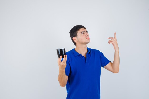 Chico joven con camiseta azul sosteniendo una taza cerca de la barbilla, levantando el dedo índice en gesto eureka y luciendo sensato, vista frontal.