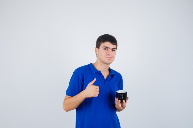 Chico joven en camiseta azul sosteniendo la taza, mostrando el pulgar hacia arriba y mirando feliz, vista frontal.