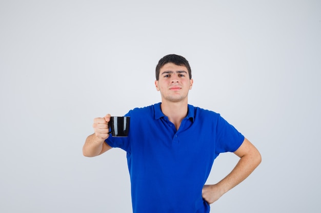 Chico joven en camiseta azul sosteniendo la taza, poniendo la mano en la cintura y mirando confiado, vista frontal.
