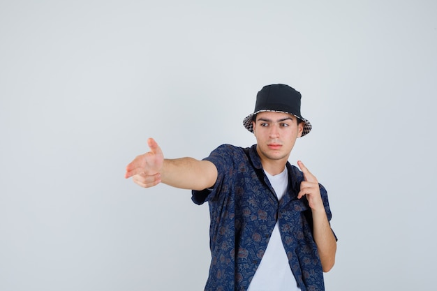 Foto gratuita chico joven con camiseta blanca, camisa floral, gorra mostrando gestos de pistola y mirando confiado