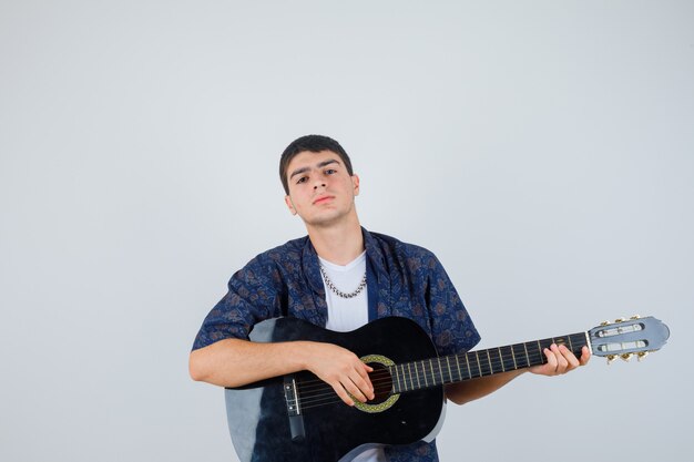 Chico joven en camiseta jugando guiter sentado aganist y mirando confiado, vista frontal.