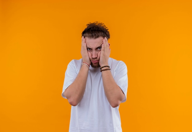 Chico joven cansado con camiseta blanca puso sus manos en las mejillas en la pared naranja aislada