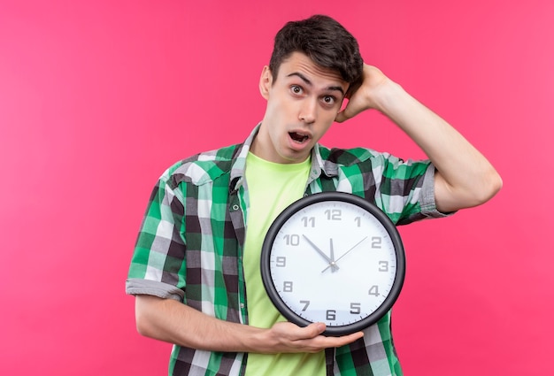 Chico joven caucásico sorprendido con camisa verde sosteniendo el reloj de pared puso su mano sobre la cabeza sobre fondo rosa aislado