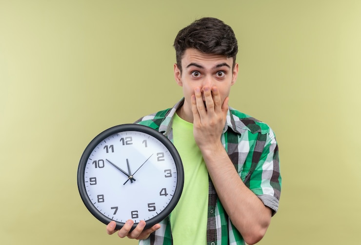 Foto gratuito chico joven caucásico sorprendido vistiendo camisa verde sosteniendo el reloj de pared con la boca cubierta con la mano sobre fondo verde aislado